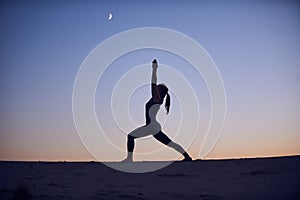 Beautiful young woman practices yoga asana Virabhadrasana 1 - warrior pose 1 in the desert at night