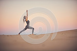 Beautiful young woman practices yoga asana Virabhadrasana 1 - warrior pose 1 in the desert at sunset