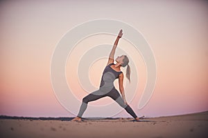 Beautiful young woman practices yoga asana Virabhadrasana 2 - warrior pose 2 in the desert at sunset