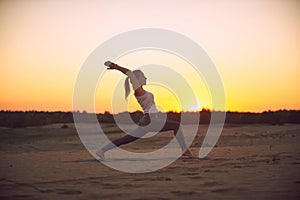 Beautiful young woman practices yoga asana Virabhadrasana 1 - warrior pose 1 in the desert at sunset