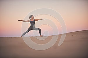 Beautiful young woman practices yoga asana Virabhadrasana 2 - warrior pose 2 in the desert at sunset