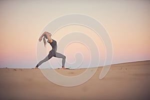 Beautiful young woman practices yoga asana Virabhadrasana 1 - warrior pose 1 in the desert at sunset