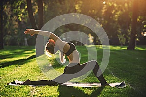 Beautiful young woman practices yoga asana Virabhadrasana 1 - warrior pose 1 in the park at sunset