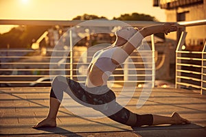 Beautiful young woman practices yoga asana Virabhadrasana 1 - warrior pose 1 in the terrace at sunset