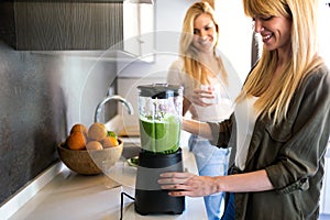 Beautiful young woman preparing detox juice in the blender and talking with friend at home.