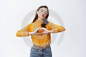 Beautiful young woman pucker lips for kiss and showing heart sign over chest, love you gesture, standing against white