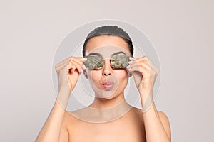 Beautiful young woman putting green tea bags on eyes against light grey background