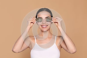Beautiful young woman putting green tea bags on skin around eyes against beige background