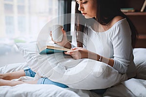 Beautiful young woman reading book and drinking coffee in bed in