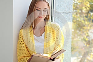 Beautiful young woman reading book near window at home