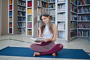 Beautiful young woman reading book and practices yoga asana Sukhasana - The Easy Sitting crosslegged Pose in the library