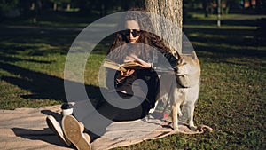 Beautiful young woman is reading book sitting on blanket under tree in park and smiling while her pedigree dog is