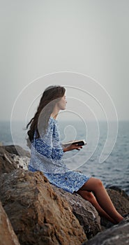 A beautiful young woman is reading a book, sitting on a large stone on seashore