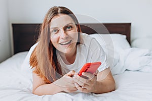 Beautiful young woman reading text message on her mobile phone in the Bed. Check social networks, send sms. The girl is