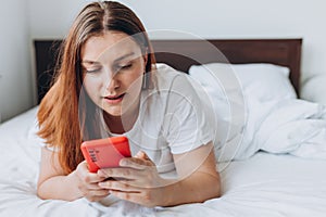 Beautiful young woman reading text message on her mobile phone in the Bed. Check social networks, send sms. The girl is