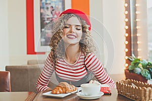 Beautiful young woman in red beret having a french breakfast with coffee and croissant, sitting at the cafe and smiling