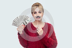 Beautiful young woman in red blouse standing, demonstrate fan of money, holding manlike mustache sticker, looking at camera with photo