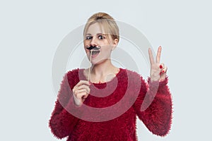 Beautiful young woman in red blouse standing, holding manlike mustache sticker, showing peace sign with two fingers and opened photo