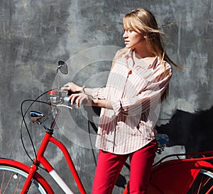 Beautiful young woman in red chino holding hands on her red vintage bicycle, standing against gray wall. Outdoors