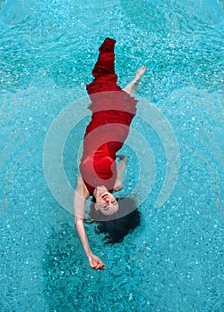 Beautiful young woman in red evening dress floating weightlessly in the pool in turquoise water