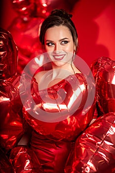 Beautiful young woman in red evening dress posing over red background with big heart shape balloons.