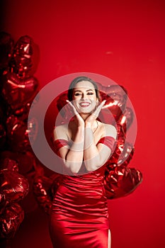 Beautiful young woman in red evening dress posing over red background with big heart shape balloons.