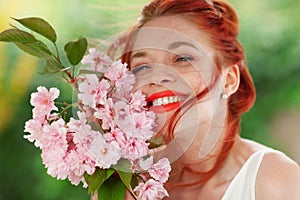 Beautiful young woman with red hair having fun standing in the garden with cherry blossom branch