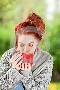 Beautiful young woman with red hair sitting in the garden, relaxing, drinking coffee