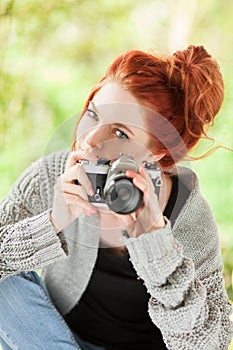 Beautiful young woman with red hair sitting in the garden taking pictures with camera