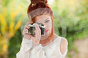 Beautiful young woman with red hair sitting in the garden taking pictures with camera