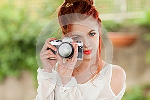 Beautiful young woman with red hair sitting in the garden taking pictures with camera