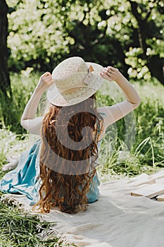 Beautiful young woman with red hair in straw hat and blue dress sitting on the plaid in the park.