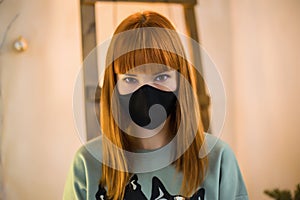 A beautiful young woman with red hair wearing a black cloth protective mask against a light colored wall with a wooden staircase