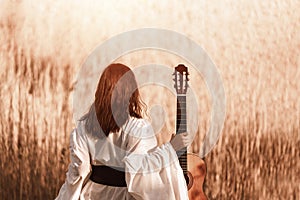 Beautiful young woman with red hair in a white medieval dress holding guitar and walking through the sunny field at sunset