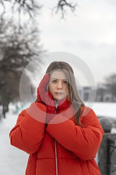 Beautiful young woman in red jacket and knitted mittens sadness look in winter park. Winter portrait of girl