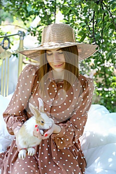 Beautiful young woman with a red manicure in a straw hat and a brown polka dot dress is sitting on a bed in a dacha and holding