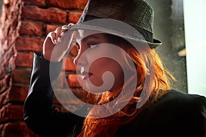 Beautiful young woman with redhair and black hat being near red brick wall inside