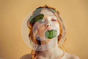 Beautiful young woman redhead blue eyes with green leaves on face. Nature organic cosmetics and makeup