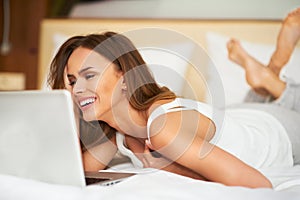 Beautiful young woman relaxing on her bed in white casual shirt using laptop