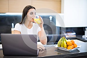 Beautiful young woman relaxing with her laptop while holding a glass of orange juice in the kitchen
