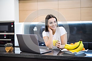 Beautiful young woman relaxing with her laptop while holding a glass of orange juice in the kitchen