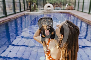 beautiful young woman relaxing in an indoor swimming pool. Cute small sausage dog on her shoulder. Summer time and lifestyle