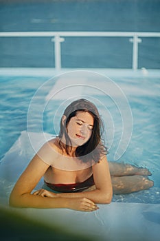 Beautiful young woman relaxing in pool, enjoying warm sunlight and wind. Brunette girl on summer vacation in luxury resort,