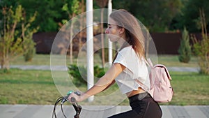 Beautiful young woman rides a bicycle in a city park on a sunny summer day. Cyclist girl with a pink backpack on her back.