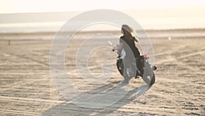 Beautiful young woman riding an old cafe racer motorcycle on desert at sunset or sunrise. Female biker. Slow motion