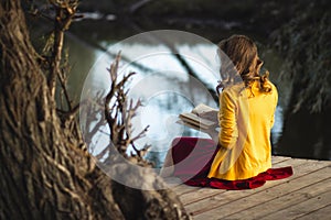 Beautiful young woman on the river banks sitting on a wooden platform with a book, teenager girl reading fiction literature on