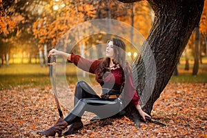 A beautiful young woman with a saber and a pistol in her hands sits near a tree in the autumn forest