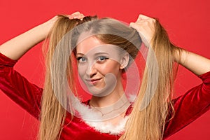 Beautiful young woman in Santa costume playing with a strand of her long silky hair, looking at camera on red background