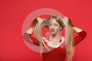 Beautiful young woman in Santa costume playing with a strand of her long silky hair, looking at camera on red background
