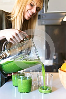 Beautiful young woman serving detox green juice into glasses at home.